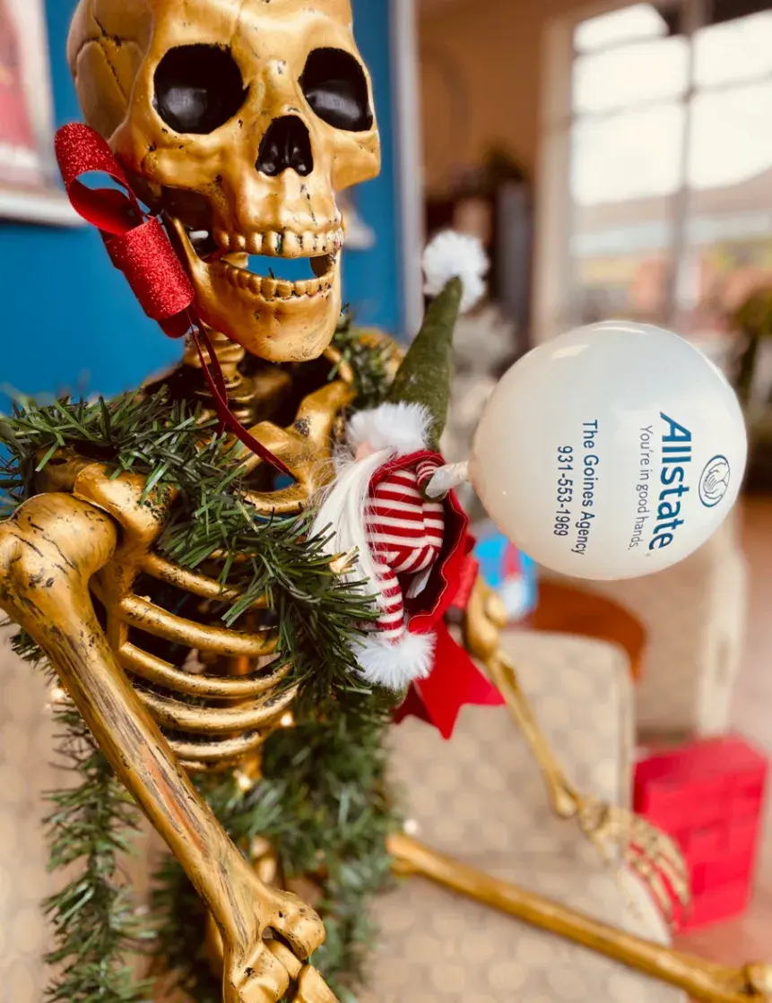 Close view of a skeleton holding a white Allstate ornament beside green garland and a holiday gnome