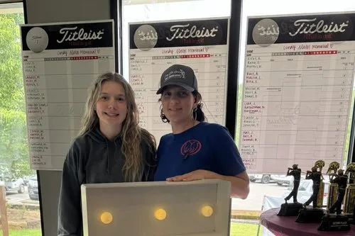Two young women stand in front of scoreboards at a golf event, smiling. One wears a black hoodie, the other a blue T-shirt and a baseball cap. Trophies are displayed on a table to the right.