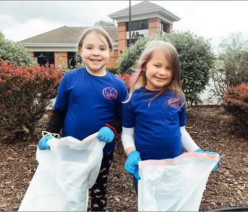 Two young girls wearing blue shirts and gloves picking up trash with trash bags outside in front of a building.